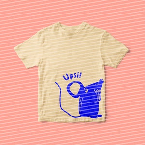 Pu&ograve; includere: Una maglietta beige chiaro con un topo di cartone animato blu e la parola "Upsi!" stampata sul davanti.