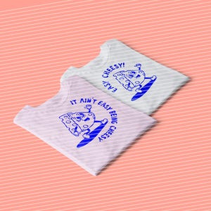 Op de afbeelding: Twee witte T-shirts met een blauwe kaas afbeelding en tekst die "It ain't easy being cheesy" en "Easy cheesy!" zegt