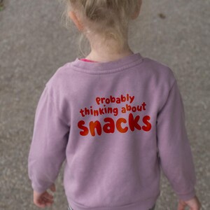 May include: A child's lavender sweatshirt with the text "Probably thinking about snacks" in red and orange. The child is wearing black and white striped trousers.