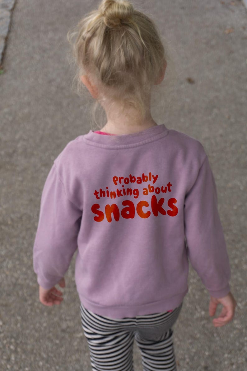 May include: A child's lavender sweatshirt with the text "Probably thinking about snacks" in red and orange. The child is wearing black and white striped trousers.