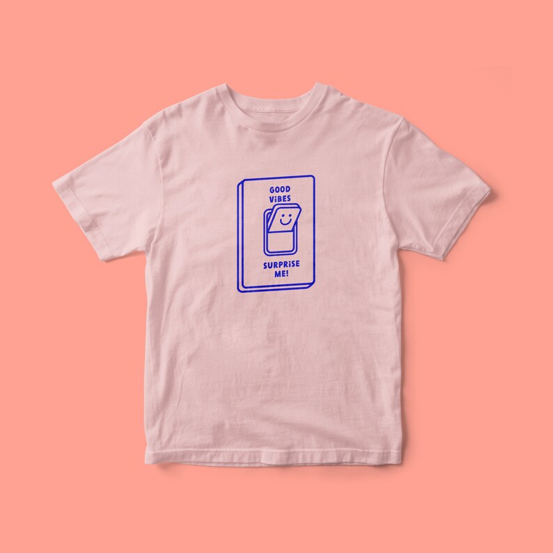 Op de afbeelding: Een roze T-shirt met een blauwe afbeelding van een lichtschakelaar met de tekst "GOOD VIBES SURPRISE ME!"