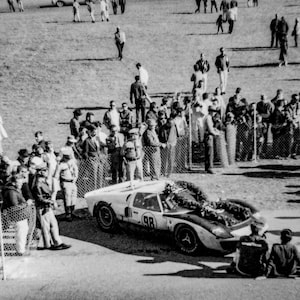 May include: Black and white photograph of a vintage race car with the number 98, adorned with a wreath, surrounded by a crowd of people. The car is parked on a paved area, with spectators and a grassy field in the background.