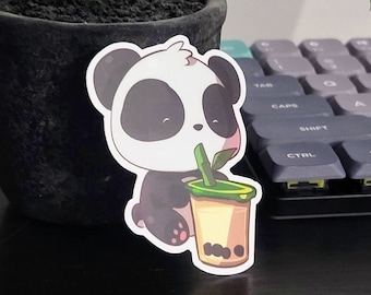 Könnte beinhalten: Ein Aufkleber mit einem schwarz-weißen Pandabären mit einem niedlichen Ausdruck, der ein grünes und braunes Boba-Tee mit einem Strohhalm hält. Der Aufkleber befindet sich auf einer schwarzen Oberfläche.