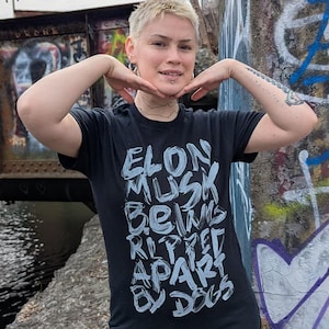 Puede incluir: Camiseta negra con el texto blanco "ELON MUSK BEING RIPPED APART BY DOGS". La persona lleva una falda negra y pantalones negros. El fondo incluye un puente y muros cubiertos de grafitis.