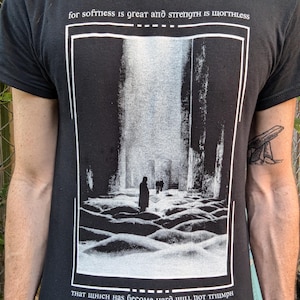 May include: Black t-shirt featuring a grayscale image of a person in a stark landscape. The design is framed with a white border and includes the text "for softness is great and strength is worthless" and "that which has become hard will not triumph."