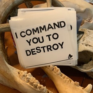 Könnte beinhalten: Ein Stapel weißer quadratischer Aufkleber mit dem schwarzen Text "I COMMAND YOU TO DESTROY". Ein kleines schwarzes Herz-Symbol befindet sich in der Ecke. Die Aufkleber liegen auf einer Holzoberfläche mit Tierknochen im Hintergrund.