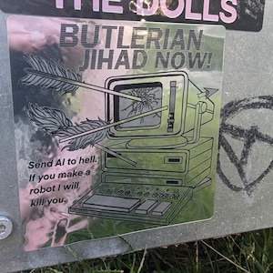 May include: A sticker with the text "THE DOLLS" in pink above the text "BUTLERIAN JIHAD NOW!" in black. The sticker features a retro computer with arrows piercing the screen. The text "Send AI to hell. If you make a robot I will kill you." is also present.