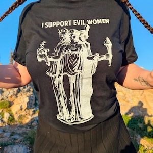 Puede incluir: Camiseta negra con el texto "I SUPPORT EVIL WOMEN" sobre un gráfico blanco de una figura de tres caras que sostiene objetos. La camiseta es de cuello redondo.