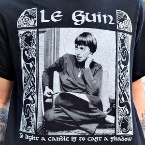 May include: Black t-shirt featuring a black and white image of a person with the text "Le Guin" above the image. The shirt also includes the quote "To light a candle is to cast a shadow."