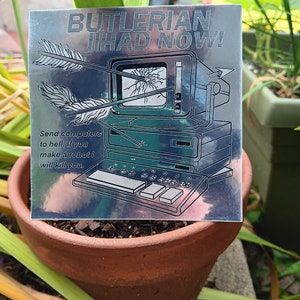 Puede incluir: Una pegatina plateada con un gráfico de ordenador retro atravesado por flechas. El texto dice "BUTLERIAN JIHAD NOW!" y "Send computers to hell. If you make a robot I will kill you."