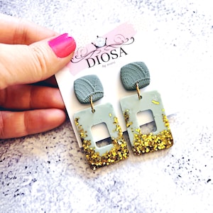 May include: A pair of green and gold square earrings with a smaller green square charm hanging from each earring. The earrings are made of polymer clay and have a glittery finish.