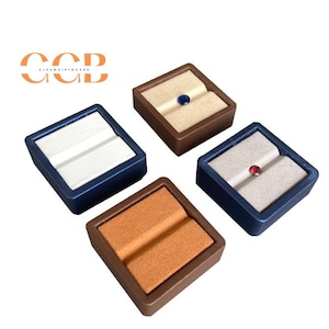 May include: Four square ring display boxes with velvet interiors. Two boxes are brown, one is blue, and one is white. Each box has a small gemstone in the center.