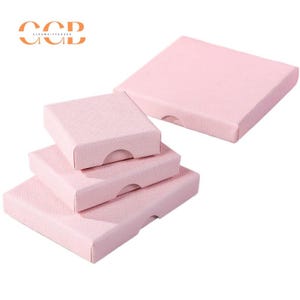 May include: A collection of pink square gift boxes of varying sizes. The boxes are made of textured paper and have a small curved cutout on one side for easy opening. The boxes are arranged in a stack and scattered on a white background.