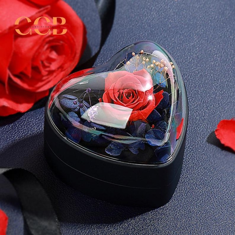 May include: A single red rose preserved in a clear heart-shaped glass dome, surrounded by blue and purple dried flowers. The dome sits on a black heart-shaped box.