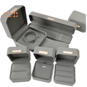 May include: A collection of grey velvet jewellery boxes featuring a rectangular shape with a small metallic gold-toned accent on the lid. The set includes various sizes for rings, bracelets, and necklaces, all lined with a soft grey interior fabric.
