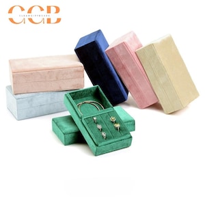 May include: A collection of rectangular jewelry boxes in various colors, including pink, blue, green, and beige. One green box is open, displaying rings and a bracelet. The boxes are made of a soft, velvet-like material.