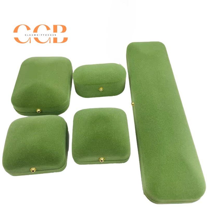 May include: A collection of five green velvet jewelry boxes in various shapes and sizes. The boxes include a rectangular, oval, and square shape, each with a gold-colored clasp. The boxes are arranged on a white background.
