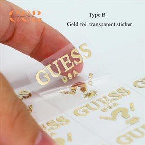 May include: Gold foil transparent stickers with the words "GUESS" and "USA" printed on them. The stickers are on a white background.