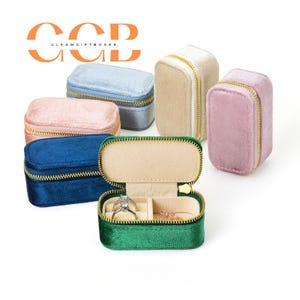 May include: A collection of small rectangular velvet jewellery boxes in various colours including blue, pink, beige, and green, each featuring a gold-tone zip closure. One green box is open, revealing a beige interior with ring slots and a small chain.