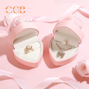 May include: Two pink heart-shaped boxes with lids open. One box contains a gold ring with a pearl and the other contains a gold necklace with a bow pendant. Both boxes are decorated with a white flower.