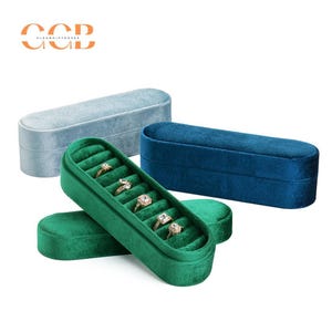 Puede incluir: Un conjunto de tres cajas de anillos de terciopelo en azul, verde y azul claro. La caja verde tiene un soporte para anillos con seis ranuras para anillos.