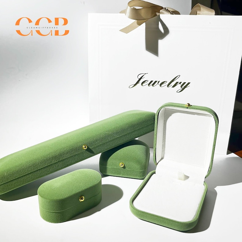 May include: An assortment of green velvet jewelry boxes in various shapes and sizes, including a long rectangular box, a square box, and an open ring box. A white gift bag with the word "Jewelry" is in the background.