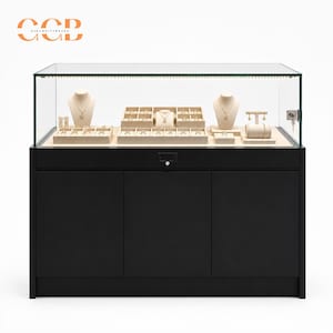 Könnte beinhalten: Eine schwarze Schmuckvitrine mit Glasoberseite und integrierter Beleuchtung. Das Innere ist mit beigefarbenen Samteinsätzen ausgestattet, darunter zwei Halskettenbüsten, ein T-förmiger Ohrringständer und mehrere Ringfächer mit einem Schloss.