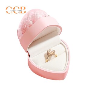 May include: A pink heart-shaped ring box with a clear dome top filled with dried flowers. The box is open and a gold ring with a pearl is inside.