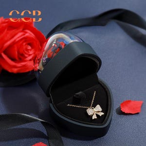 May include: A black heart-shaped box with a gold necklace with a sparkling bow pendant inside. The box is open and the necklace is visible. The box is sitting on a blue surface with a red rose and black ribbon in the background.