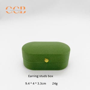 May include: A green, oval-shaped earring studs box with a gold-colored clasp. The box is made of a soft, textured material. The text "Earring studs box" is below the box. Dimensions are 9.4 x 4 x 3.3 cm, and it weighs 24g.