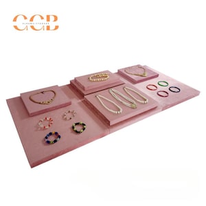 May include: A display of jewelry on pink platforms. The jewelry includes necklaces, bracelets, and rings. The necklaces feature pearls and gold chains. The bracelets are made of colorful beads. The rings are in various colors. The display is set against a white background.
