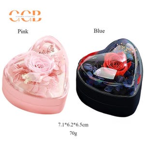 May include: Two heart-shaped boxes with lids, one pink and one black, each containing a preserved flower arrangement. The pink box has a pink rose and white flowers, while the black box has a red rose and blue flowers. The boxes are 7.1 inches by 6.2 inches by 6.5 inches and weigh 70 grams.
