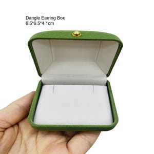 May include: Open green velvet dangle earring box with a white interior. The box is rectangular, with a gold-colored clasp. Text on the image reads: Dangle Earring Box 6.5*6.5*4.1cm.