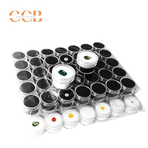 Può includere: Una collezione di piccoli contenitori di plastica trasparente con coperchi neri, ciascuno contenente una gemma. Le gemme sono di vari colori, tra cui verde, giallo, rosso e trasparente. I contenitori sono disposti su un foglio di plastica trasparente.