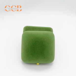 May include: A green, square-shaped jewelry box with a textured, velvety surface. The box has a hinged lid and a small, yellow accent at the base. The logo "CCB CLEAN GIFT BOXES" is in the upper left corner.