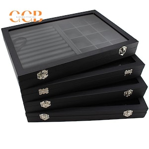 May include: A stack of four black wooden jewelry display boxes with glass tops. Each box has a silver clasp and a divided interior for organizing rings, necklaces, and other accessories. The top box has the logo "CCB" in orange.