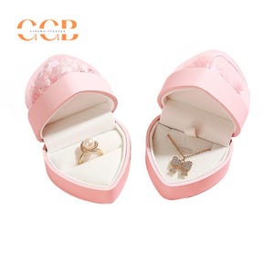 May include: Two pink heart-shaped jewelry boxes with lids open. The box on the left has a ring with a pearl in the center. The box on the right has a necklace with a bow pendant.
