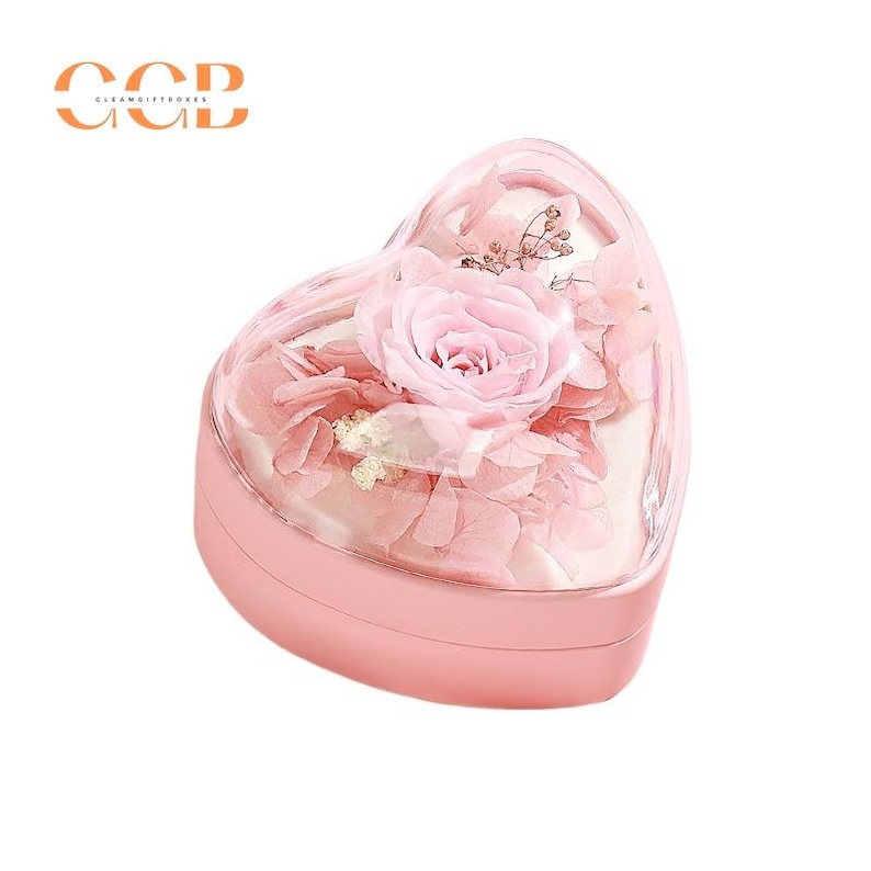 May include: A pink heart-shaped box with a clear lid containing a preserved pink rose and other flowers. The box is made of plastic and has a smooth, glossy finish.