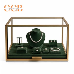 Pode incluir: Um expositor de joias retangular com moldura de metal dourado e painéis de vidro, apresentando um interior de veludo verde escuro. No interior, um busto de veludo verde exibe um colar de pérolas, brincos, anéis e uma pulseira a condizer.