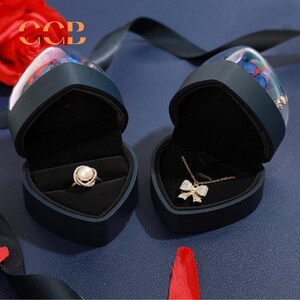 May include: Two heart-shaped black velvet jewelry boxes with clear lids. The boxes are open and contain a ring with a pearl and a necklace with a bow pendant. The boxes are decorated with a floral design.