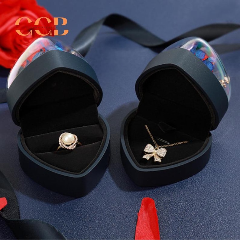 May include: Two heart-shaped black velvet jewelry boxes with clear lids. The boxes are open and contain a ring with a pearl and a necklace with a bow pendant. The boxes are decorated with a floral design.