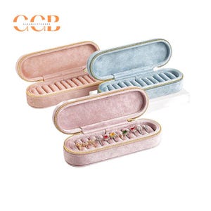 May include: Two pink and one blue velvet ring boxes with gold zips. The boxes are open and show rows of ring slots. The pink box has colourful rings inside.