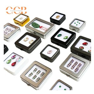 K&ouml;nnte beinhalten: Eine Sammlung kleiner Ausstellungsboxen mit verschiedenen Edelsteinen und dekorativen Gegenst&auml;nden. Die Boxen sind in verschiedenen Farben erh&auml;ltlich, darunter Schwarz, Silber und Gold, mit transparenten Deckeln. Der Inhalt umfasst farbige Edelsteine und Perlen.