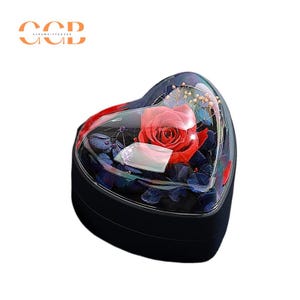 May include: A heart-shaped box with a clear lid, containing a preserved red rose and other dried flowers. The box is black and has a glossy finish.