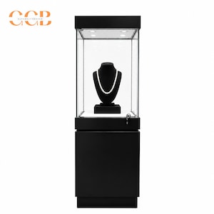 Könnte beinhalten: Eine hohe, schwarze, rechteckige Vitrine mit Glaspaneelen im oberen Bereich. Im Inneren präsentiert eine schwarze Büste eine weiße Perlenkette. Der Sockel ist massiv schwarz und verfügt über ein Schloss an der Vorderseite. Der obere Innenraum ist beleuchtet.