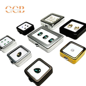 Könnte beinhalten: Eine Sammlung kleiner Ausstellungsboxen, die jeweils Edelsteine enthalten. Die Boxen sind in verschiedenen Farben erhältlich, darunter Schwarz, Silber und Gold. Die Edelsteine sind smaragdgrün, klar und perlmuttartig. Die Boxen sind quadratisch und haben eine transparente Oberseite.