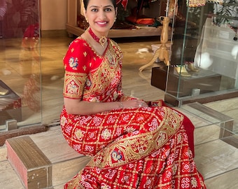 Può includere: Una donna indossa un sari rosso e oro ricamato con una blusa rossa. Il sari ha un motivo geometrico ed è decorato con paillettes e perline dorate. La donna è seduta su una scalinata.