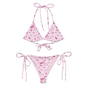 May include: A two-piece bikini set in a light pink floral pattern. The top is a halter style with triangle cups and ties at the neck and back. The bottoms are a low-rise style with tie sides. The floral pattern includes small pink roses and white polka dots.
