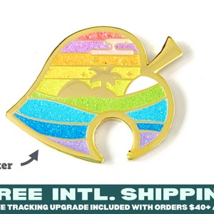 May include: Gold-coloured enamel pin shaped like a leaf with a rainbow sunset and glitter detail. The pin features a palm tree and a white crescent moon. The text "Glitter" is visible in the image.