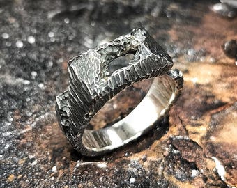 May include: A textured silver ring with a rectangular top featuring a unique, carved design. The ring's band is smooth and polished, contrasting with the rough texture of the top. The ring is displayed on a dark, textured surface.